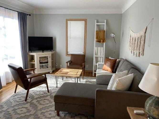 Cozy small living room with a white corner TV stand, grey sectional sofa, wooden accent chairs, patterned area rug and natural daylight